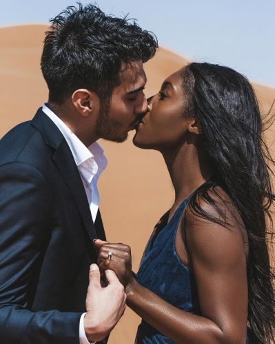 A young couple embraces lovingly with foreheads touching as they stand in the empty Dubai desert landscape, dressed elegantly.