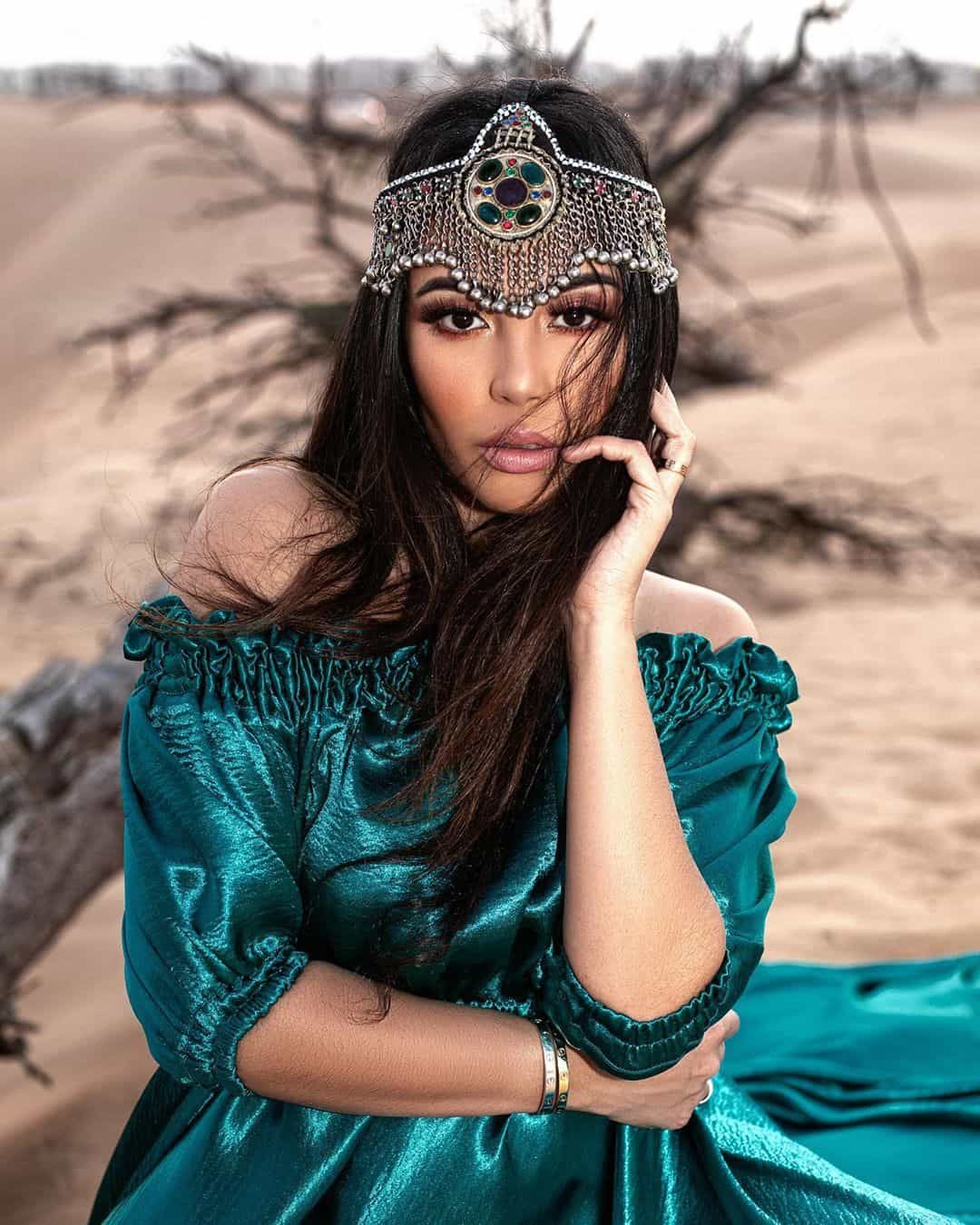 A woman wearing tiara and green dress stands in the Dubai desert, with sand dunes stretching out behind her.